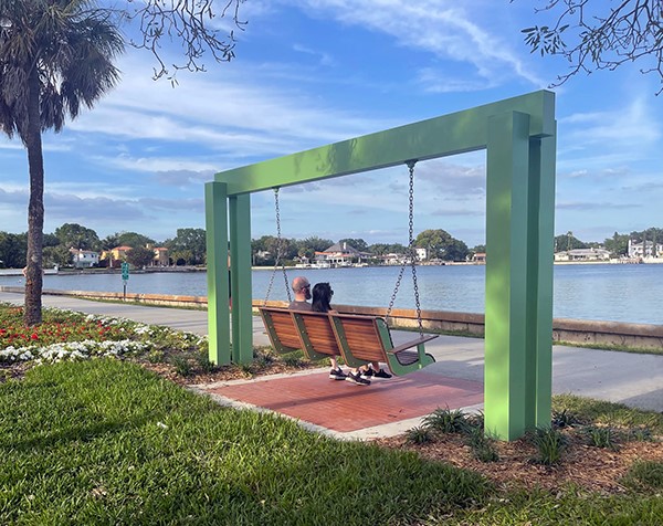An image of a couple sitting on a green bench swing in Flora Wylie Park overlooking Tampa Bay